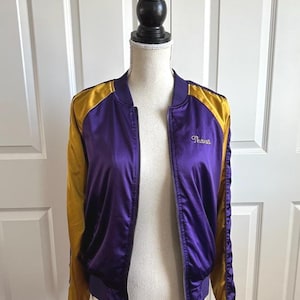 May include: Purple and gold satin bomber jacket with a zipper closure. The jacket has gold sleeves and shoulder accents, with the word "Shanes" embroidered on the chest. The jacket is displayed on a mannequin.