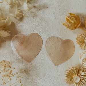 May include: Two heart-shaped, translucent pink glass coasters on a white textured background with dried flowers and other decorative items.