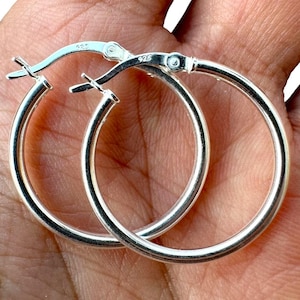 May include: A pair of shiny silver hoop earrings. The earrings are round with a hinged closure. The metal is stamped with "925". The earrings are displayed against a skin-toned background.