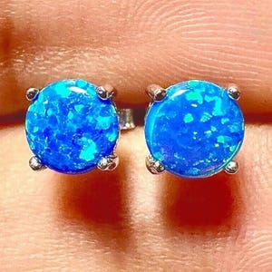 May include: A pair of round, blue opal stud earrings. Each earring features a vibrant, speckled blue stone set in a silver-colored metal with four prongs. The earrings are displayed against a blurred background.