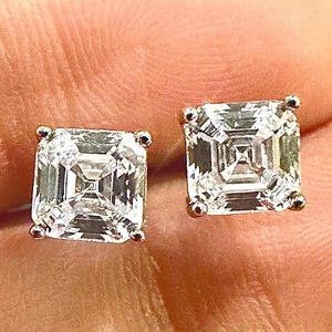 May include: A pair of square-cut diamond stud earrings. The diamonds are clear and sparkling, set in a gold-colored metal. The earrings are displayed against a neutral background.