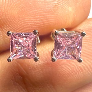 May include: A pair of square-cut, pink gemstone stud earrings. Each earring has a silver-colored setting with four small, round accents at the corners. The gemstones are a light pink color.