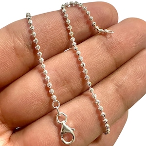 May include: A delicate silver ball chain necklace with a lobster clasp. The chain is composed of small, round, textured beads. The necklace is shown against a neutral background.