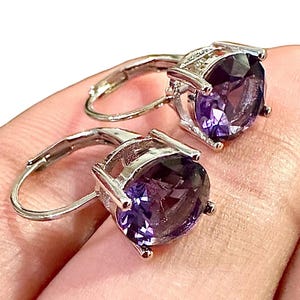 K&ouml;nnte beinhalten: Ein Paar silberfarbene Klappcreolen, jeweils mit einem gro&szlig;en, oval geschliffenen, tiefvioletten Edelstein. Die Edelsteine sind in einer Vier-Zacken-Fassung gefasst, die ihre Farbe und Brillanz hervorhebt.