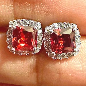 May include: A pair of square-cut, red gemstone stud earrings. Each earring is surrounded by a halo of small, clear stones, creating a sparkling border. The earrings are set in a silver-colored metal.