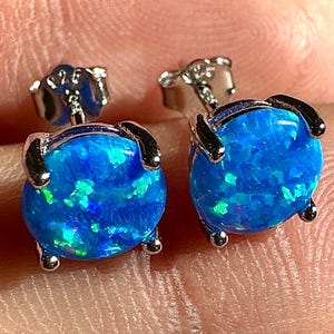 May include: A pair of stud earrings featuring vibrant blue opal stones. The opals have a round shape and are set in a silver-colored metal. The earrings are designed with a classic post-and-clutch closure.