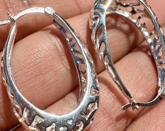 May include: A pair of silver oval hoop earrings. The earrings have a filigree design with cut-out patterns. The earrings are shown against a skin-toned background. The earrings are made of a shiny, reflective metal.