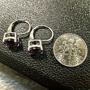 K&ouml;nnte beinhalten: Ein Paar silberne Klappb&uuml;gel-Ohrringe mit ovalen, tiefvioletten Edelsteinen. Eine US-Dime liegt neben den Ohrringen zum Gr&ouml;&szlig;envergleich. Die Ohrringe liegen auf einer strukturierten, dunklen Oberfl&auml;che.