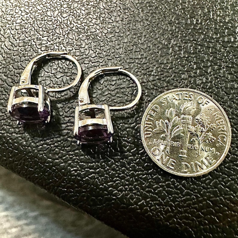K&ouml;nnte beinhalten: Ein Paar silberne Klappb&uuml;gel-Ohrringe mit ovalen, tiefvioletten Edelsteinen. Eine US-Dime liegt neben den Ohrringen zum Gr&ouml;&szlig;envergleich. Die Ohrringe liegen auf einer strukturierten, dunklen Oberfl&auml;che.