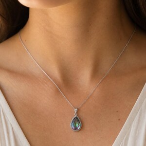 Pu&ograve; includere: Una collana d'argento con un pendente a forma di goccia. Il pendente presenta una grande pietra iridescente con un alone di piccole pietre trasparenti. La collana &egrave; indossata su un top bianco.