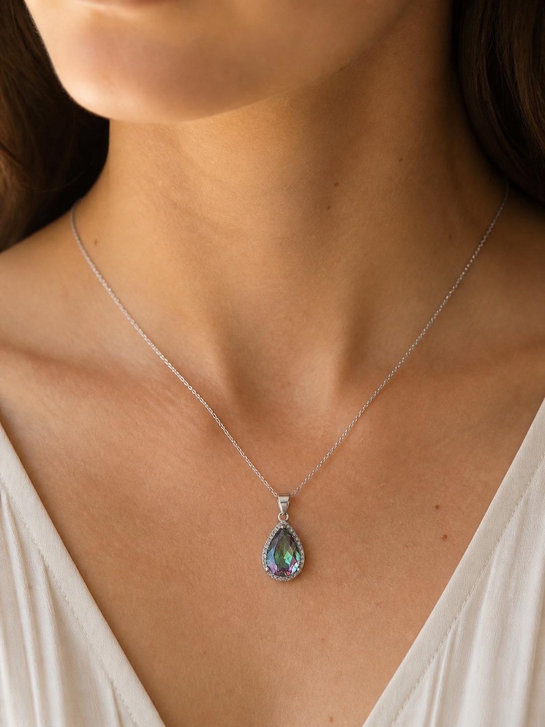 Pu&ograve; includere: Una collana d'argento con un pendente a forma di goccia. Il pendente presenta una grande pietra iridescente con un alone di piccole pietre trasparenti. La collana &egrave; indossata su un top bianco.