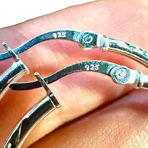 May include: A pair of silver hoop earrings featuring a hinged closure mechanism. Each earring is clearly stamped with the hallmark 925, indicating sterling silver composition. The metal surface is polished and reflective, showing the hinge detail.
