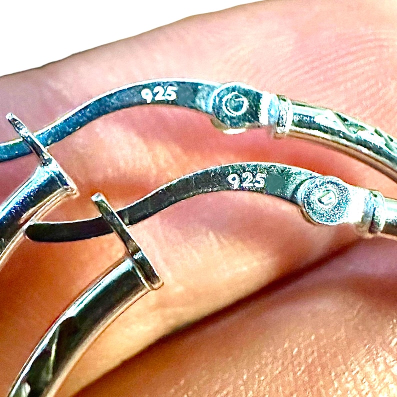 May include: A pair of silver hoop earrings featuring a hinged closure mechanism. Each earring is clearly stamped with the hallmark 925, indicating sterling silver composition. The metal surface is polished and reflective, showing the hinge detail.