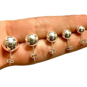 May include: Five silver ball stud earrings of varying sizes are displayed. Each earring features a polished, spherical design and a post with a butterfly clutch backing. The earrings are shown against a light background.