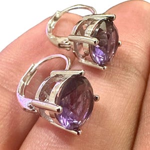 K&ouml;nnte beinhalten: Ein Paar Ohrringe mit B&uuml;gelverschluss, die gro&szlig;e, oval geschliffene, violette Edelsteine in silberfarbenem Metall aufweisen. Die Ohrringe haben ein klassisches Design mit einem sicheren B&uuml;gelverschluss.