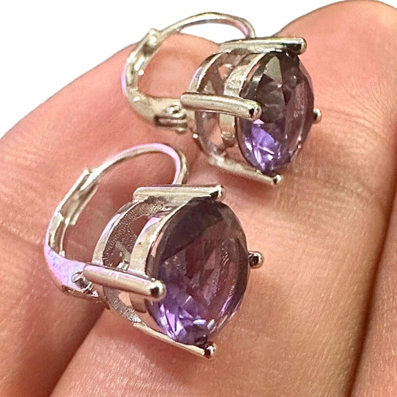 K&ouml;nnte beinhalten: Ein Paar Ohrringe mit B&uuml;gelverschluss, die gro&szlig;e, oval geschliffene, violette Edelsteine in silberfarbenem Metall aufweisen. Die Ohrringe haben ein klassisches Design mit einem sicheren B&uuml;gelverschluss.