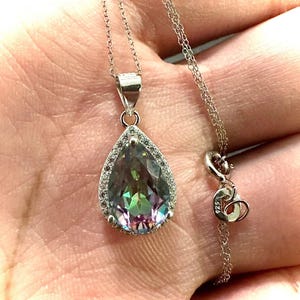 Pu&ograve; includere: Collana d'argento con un ciondolo a forma di goccia. Il ciondolo presenta una pietra preziosa iridescente, circondata da piccole pietre trasparenti. La catena e la chiusura sono in argento.