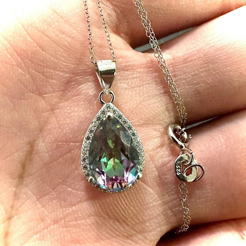Pu&ograve; includere: Collana d'argento con un ciondolo a forma di goccia. Il ciondolo presenta una pietra preziosa iridescente, circondata da piccole pietre trasparenti. La catena e la chiusura sono in argento.