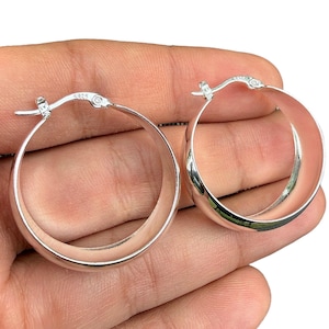 May include: A pair of shiny silver hoop earrings. The earrings are round with a smooth, polished surface. The clasp is visible on each earring. The earrings are held in a hand.