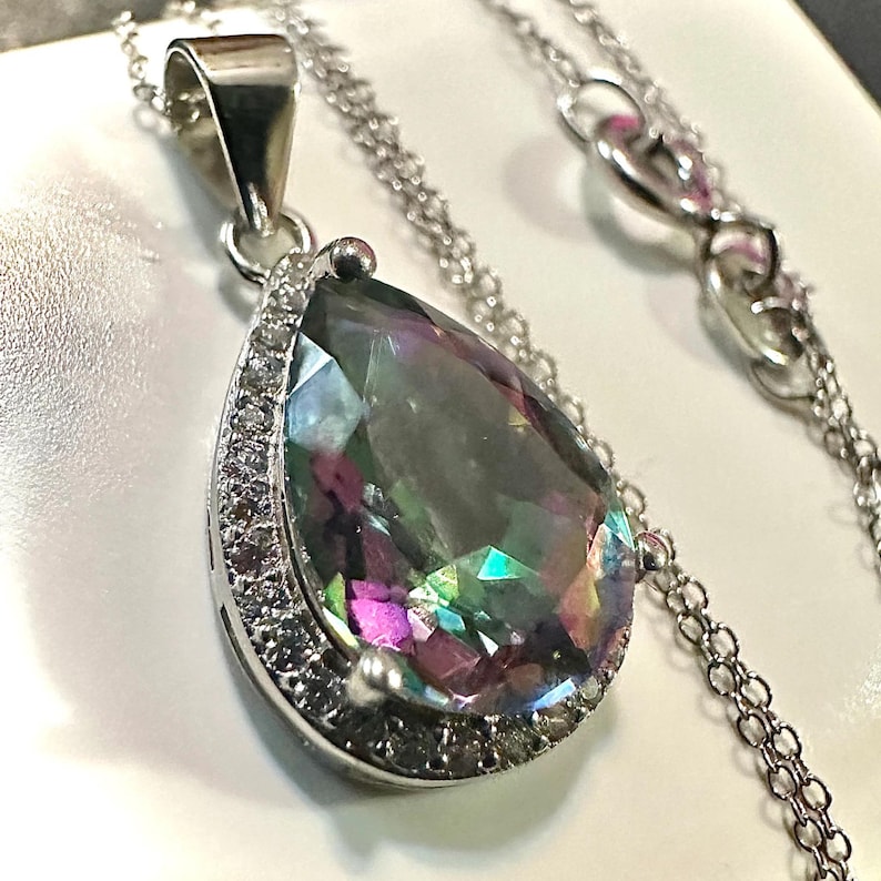 Pu&ograve; includere: Una collana d'argento con un grande pendente a forma di goccia. Il pendente presenta una pietra preziosa iridescente con un effetto arcobaleno. La pietra &egrave; circondata da piccole pietre trasparenti. La catena &egrave; delicata e argentata.