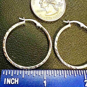 May include: A pair of silver-toned hoop earrings with a textured, etched pattern along the outer surface. The earrings are positioned above a blue ruler showing a diameter of approximately 1 inch, with a US quarter coin placed above them.