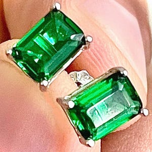 May include: A pair of emerald-cut, green gemstone stud earrings with a silver setting. The earrings have a rectangular shape and are set with four small white prongs. The gemstones have a vibrant, reflective quality.