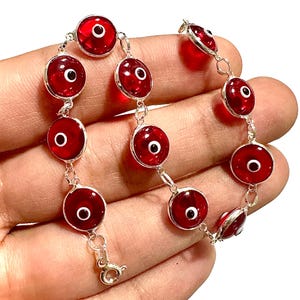 May include: A silver bracelet featuring a series of red, eye-shaped charms. Each charm has a white center and a black pupil. The bracelet is connected by small silver links and has a clasp closure.