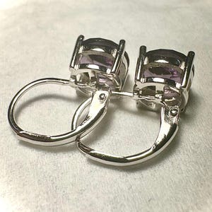 K&ouml;nnte beinhalten: Ein Paar silberne Klappb&uuml;gel-Ohrringe mit ovalen, hellvioletten Edelsteinen. Die Ohrringe sind in einem silberfarbenen Metall gefasst und werden auf einer wei&szlig;en Oberfl&auml;che pr&auml;sentiert. Die Edelsteine haben einen facettierten Schliff.