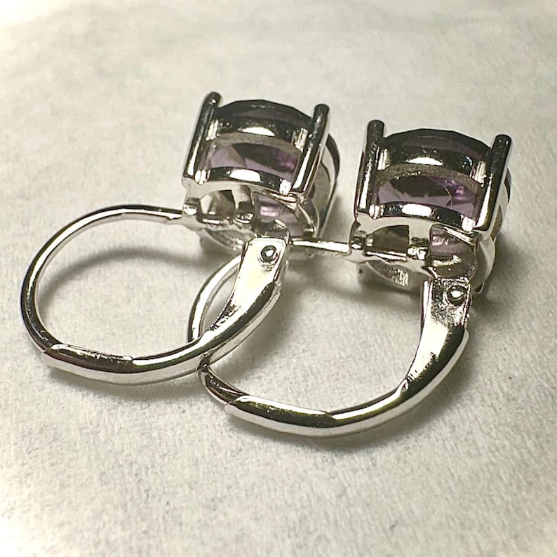 K&ouml;nnte beinhalten: Ein Paar silberne Klappb&uuml;gel-Ohrringe mit ovalen, hellvioletten Edelsteinen. Die Ohrringe sind in einem silberfarbenen Metall gefasst und werden auf einer wei&szlig;en Oberfl&auml;che pr&auml;sentiert. Die Edelsteine haben einen facettierten Schliff.