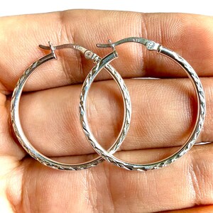 May include: A pair of sterling silver hoop earrings featuring a textured, diamond-cut pattern along the outer surface. Each hoop is secured with a hinged post closure and is clearly stamped with the 925 hallmark indicating sterling silver composition.