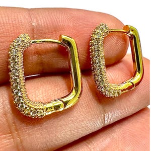 May include: A pair of gold-toned hoop earrings with a rectangular shape. The front-facing surface of each hoop is encrusted with small, clear, sparkling stones, while the inner and side surfaces feature a smooth, polished gold-toned metal finish.