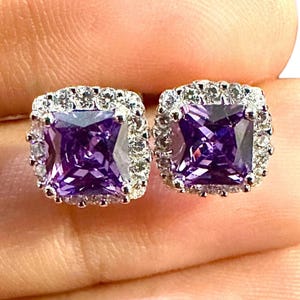 May include: A pair of square stud earrings. Each earring features a large, faceted, purple gemstone surrounded by a halo of smaller, clear stones. The earrings are set in a silver-colored metal.