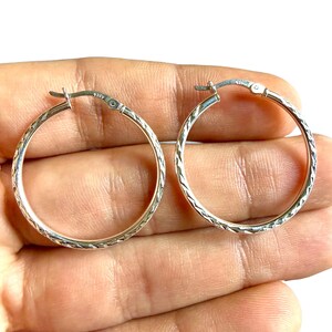 May include: A pair of silver hoop earrings featuring a textured, diamond-cut pattern along the outer surface. Each earring is constructed with a hinged post closure and is clearly stamped with the 925 hallmark, indicating sterling silver composition.