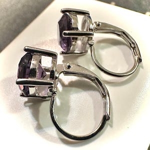 K&ouml;nnte beinhalten: Ein Paar Ohrringe mit B&uuml;gelverschluss und facettierten, oval geschliffenen lila Edelsteinen, die in silberfarbenem Metall gefasst sind. Die Ohrringe haben ein klassisches Design mit sicherem Verschluss.