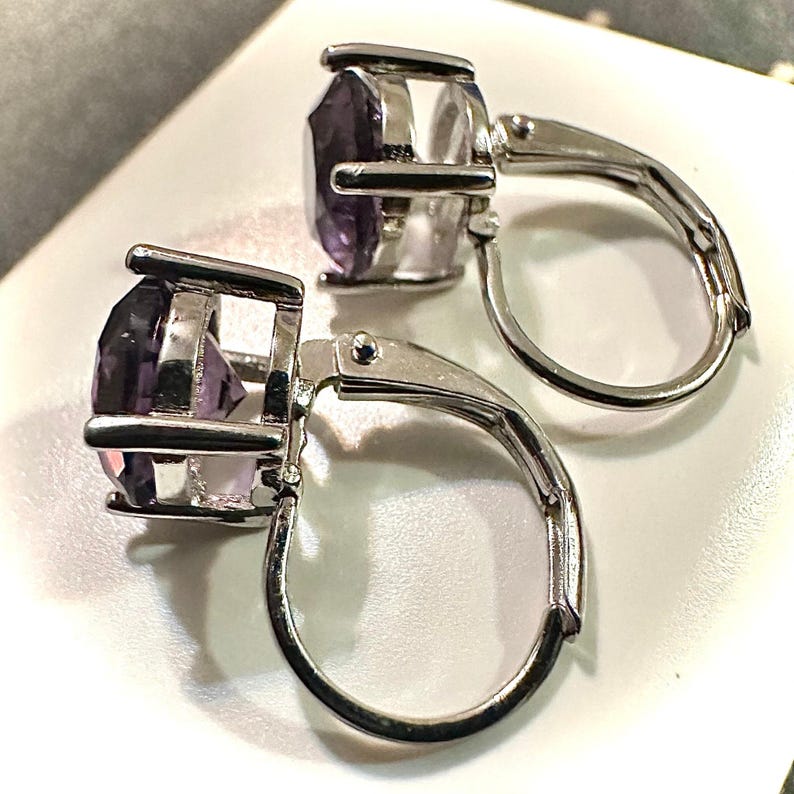 K&ouml;nnte beinhalten: Ein Paar Ohrringe mit B&uuml;gelverschluss und facettierten, oval geschliffenen lila Edelsteinen, die in silberfarbenem Metall gefasst sind. Die Ohrringe haben ein klassisches Design mit sicherem Verschluss.