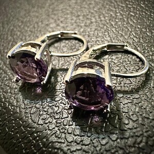 K&ouml;nnte beinhalten: Ein Paar silberfarbene Ohrringe mit B&uuml;gelverschluss, jeweils mit einem gro&szlig;en, oval geschliffenen, tiefvioletten Edelstein. Die Ohrringe sind in einem schlichten, eleganten Design gehalten, das die leuchtende Farbe der Steine hervorhebt.