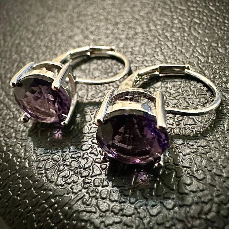 K&ouml;nnte beinhalten: Ein Paar silberfarbene Ohrringe mit B&uuml;gelverschluss, jeweils mit einem gro&szlig;en, oval geschliffenen, tiefvioletten Edelstein. Die Ohrringe sind in einem schlichten, eleganten Design gehalten, das die leuchtende Farbe der Steine hervorhebt.