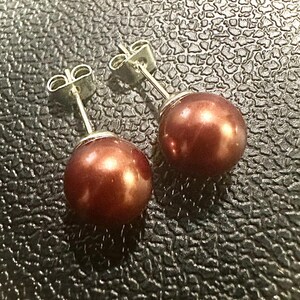 K&ouml;nnte beinhalten: Ein Paar Ohrstecker mit runden, dunkel kupferfarbenen Perlen. Jede Perle ist sicher auf einem silberfarbenen Metallstift mit passendem Schmetterlingsverschluss montiert, was ein klassisches und schlichtes Design f&uuml;r diese Schmuckst&uuml;cke ergibt.