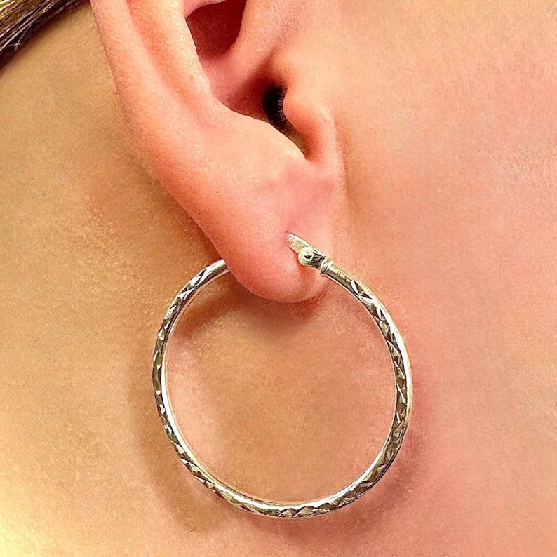 May include: A silver hoop earring featuring a textured, diamond-cut pattern along the outer surface. The circular earring has a hinged closure mechanism at the top where it connects to the earlobe, showcasing a polished metallic finish throughout.