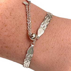 May include: A silver bracelet with a woven chain design. The bracelet features a delicate chain with a small ball and a clasp. The bracelet is shown on a person's wrist.