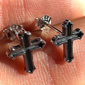 May include: A pair of silver-colored cross stud earrings. The earrings have a detailed design with a dark, textured center and a polished, raised border. The cross shape is clearly defined, and the studs are set against a blurred background.