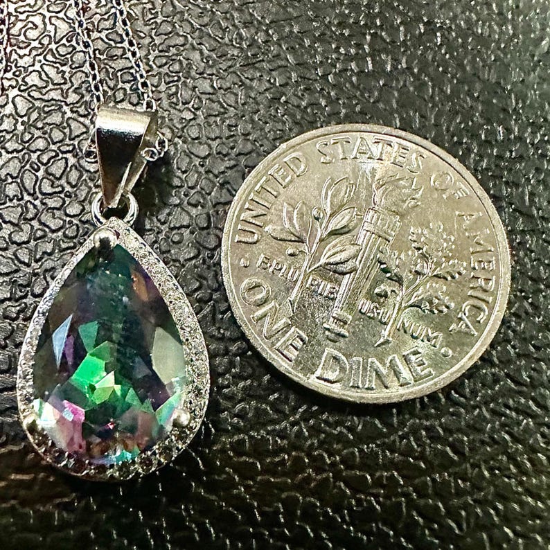 Pu&ograve; includere: Una collana con pendente in argento con una pietra preziosa iridescente a forma di pera e un alone di piccole pietre trasparenti. Una moneta da dieci centesimi americana &egrave; accanto al pendente. La pietra preziosa mostra sfumature di verde, viola e blu.