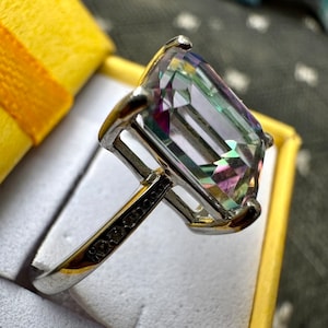 May include: A silver ring with a large, emerald-cut, iridescent gemstone. The gemstone has a rainbow of colors, including pink, purple, green, and blue.
