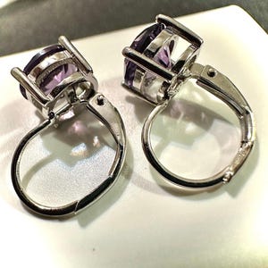 K&ouml;nnte beinhalten: Ein Paar Ohrringe mit Klappb&uuml;geln aus silberfarbenem Metall, jeweils mit einem gro&szlig;en, oval geschliffenen, violetten Edelstein. Die Ohrringe werden auf einer wei&szlig;en Oberfl&auml;che pr&auml;sentiert, um die Edelsteine hervorzuheben.