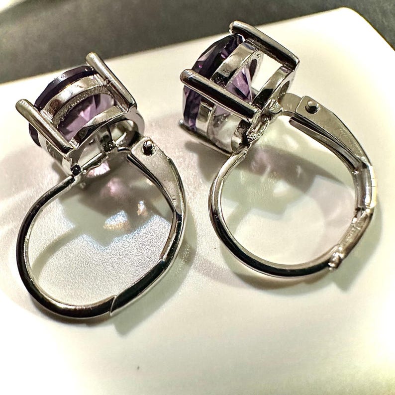 K&ouml;nnte beinhalten: Ein Paar Ohrringe mit Klappb&uuml;geln aus silberfarbenem Metall, jeweils mit einem gro&szlig;en, oval geschliffenen, violetten Edelstein. Die Ohrringe werden auf einer wei&szlig;en Oberfl&auml;che pr&auml;sentiert, um die Edelsteine hervorzuheben.