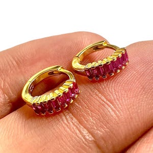 May include: A pair of gold-colored hoop earrings, each adorned with a row of rectangular, vibrant pink gemstones. The earrings are shown against a skin-toned background, highlighting their color and design.