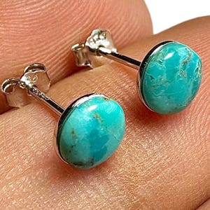May include: A pair of turquoise stud earrings. Each earring features a round, polished turquoise stone set in a silver-colored bezel. The turquoise stones have a vibrant blue-green color with natural variations. The earrings have a post and butterfly backing.