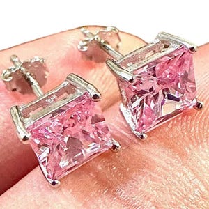 May include: A pair of square-cut pink gemstone stud earrings with a silver-colored setting. The earrings are displayed on a light pink surface, showcasing their sparkling facets. The studs have a classic, elegant design.