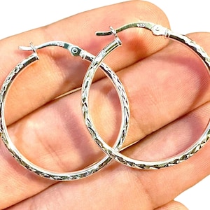 May include: A pair of sterling silver hoop earrings featuring a textured, diamond-cut pattern along the outer surface. Each hoop is secured with a hinged post closure and is clearly stamped with the 925 hallmark indicating sterling silver composition.