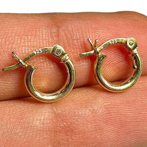 May include: A pair of small, gold-colored hoop earrings. Each earring has a hinged closure and is marked with "925". The earrings are round and have a polished finish.